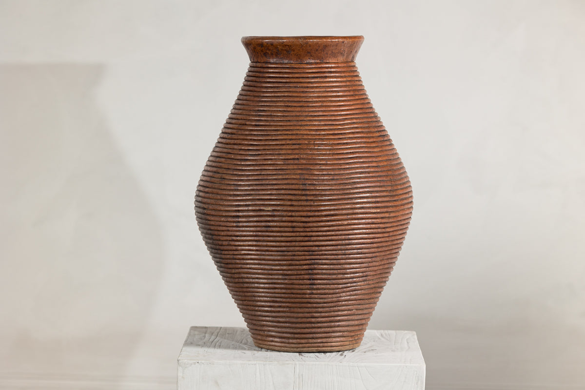 Brown ribbed pottery vase with burnished surface on white pedestal, light gray background, close-up.