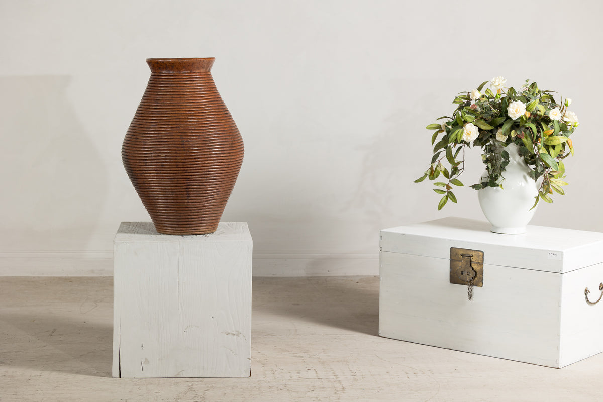 Brown ribbed pottery vase with burnished surface displayed on a white pedestal beside a trunk with florals.