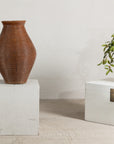 Brown ribbed pottery vase with burnished surface displayed on a white pedestal beside a trunk with florals.