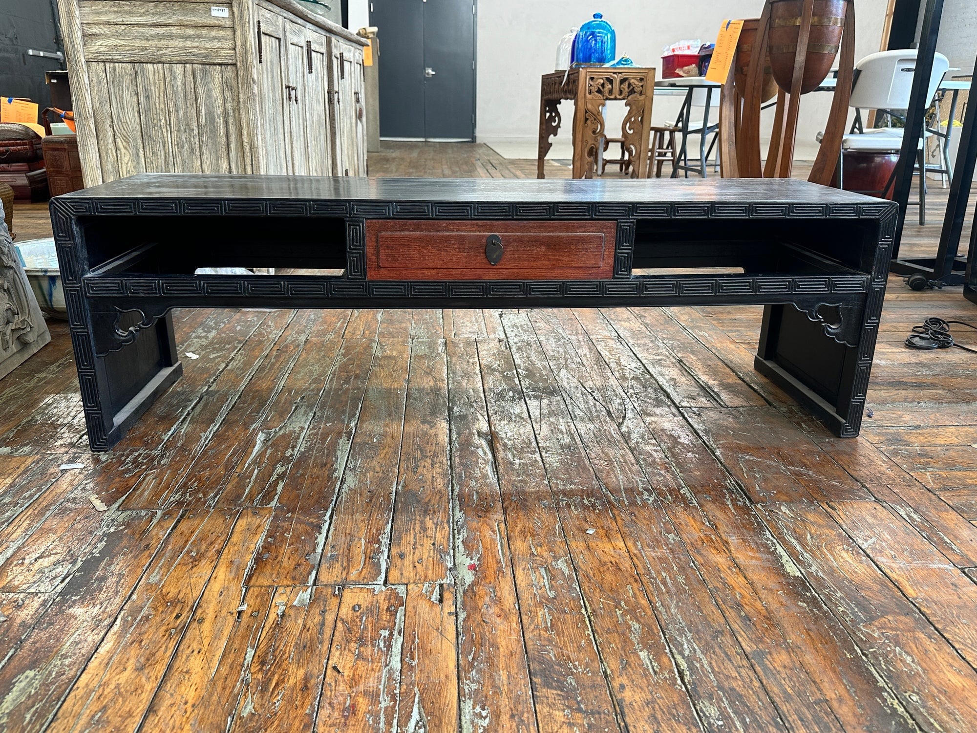 Black lacquer patina carved wood coffee table with red drawer and open shelves on rustic wood floor.