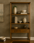 Tall wooden three-shelf bookcase with two drawers, styled with beige vases, bowls, and books beside painting and round table.