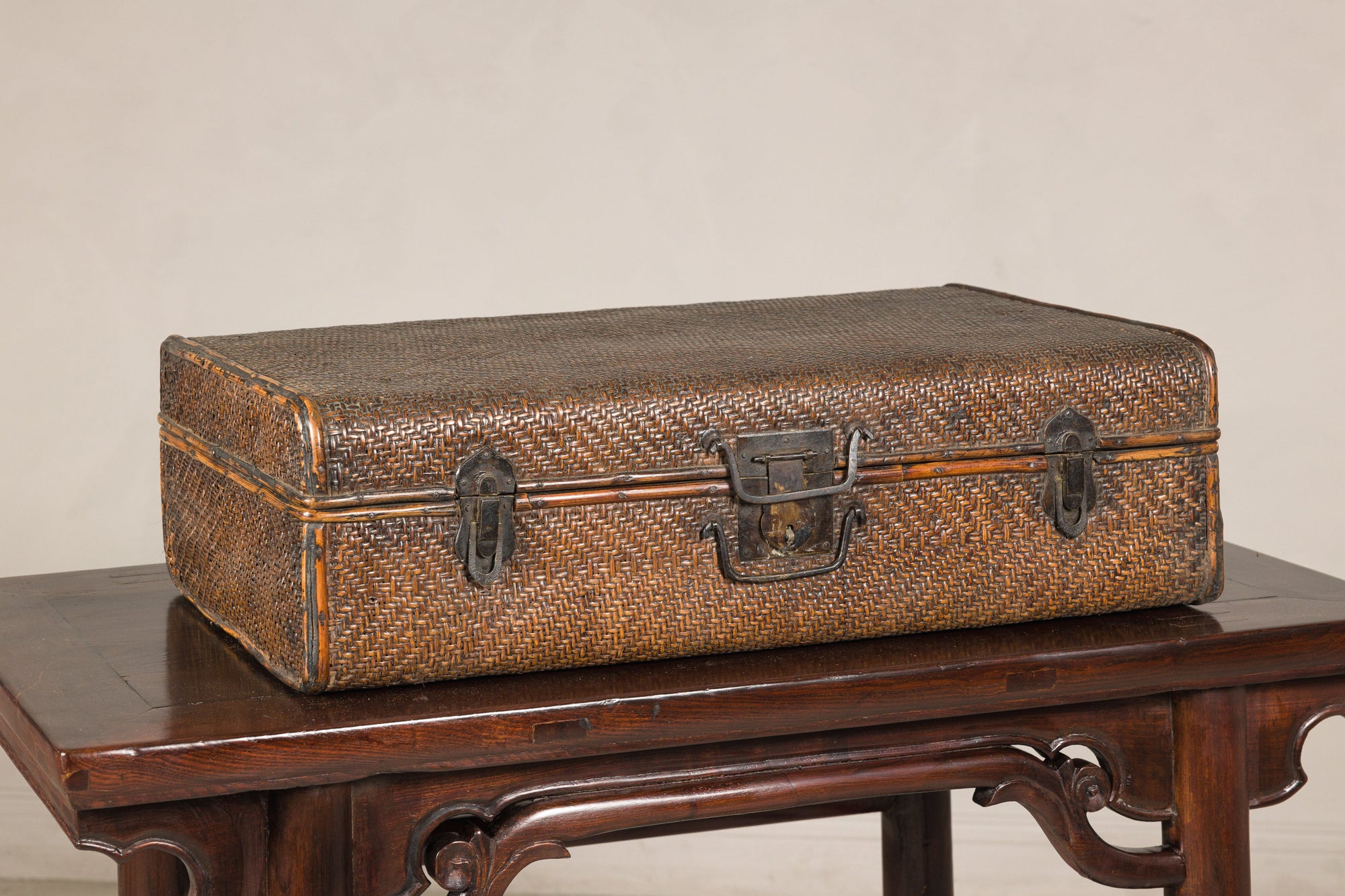Brown rattan and iron Qing Dynasty suitcase trunk on dark wooden table, plain backdrop, close-up.