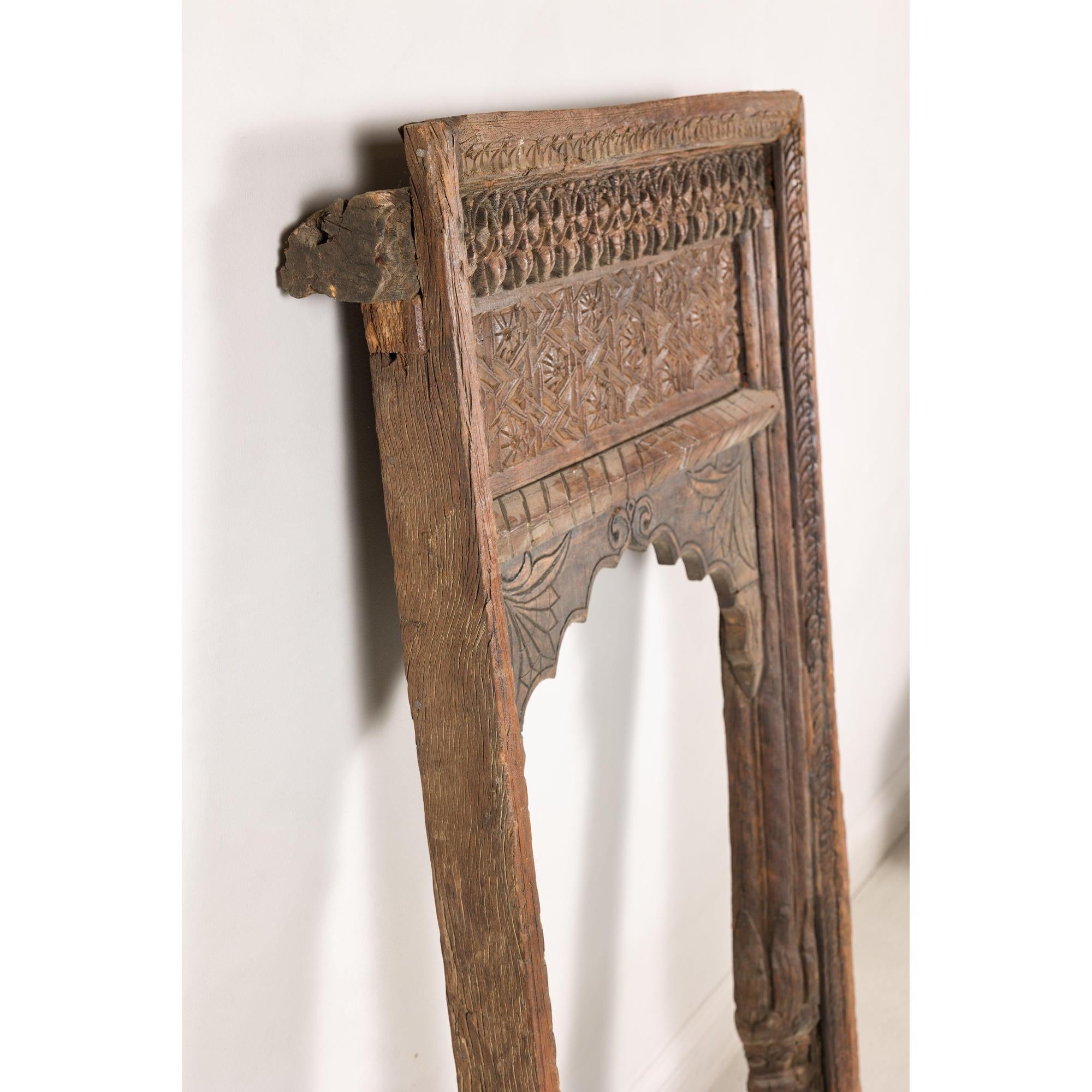 Brown wooden hand-carved Indian architectural frame with arched opening leaning against a white textured wall.