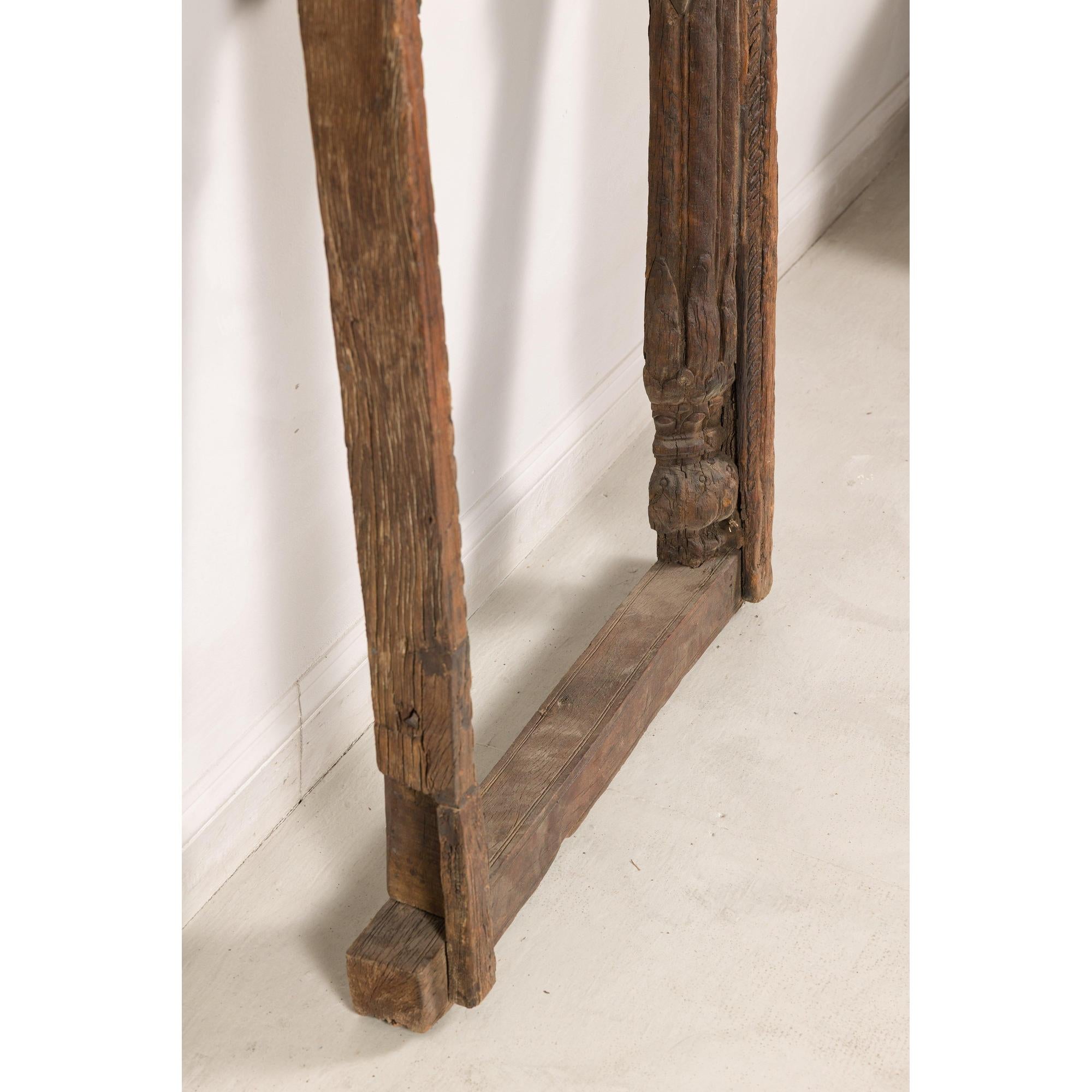 Brown wood hand-carved Indian frame with arched opening on light floor against white wall.