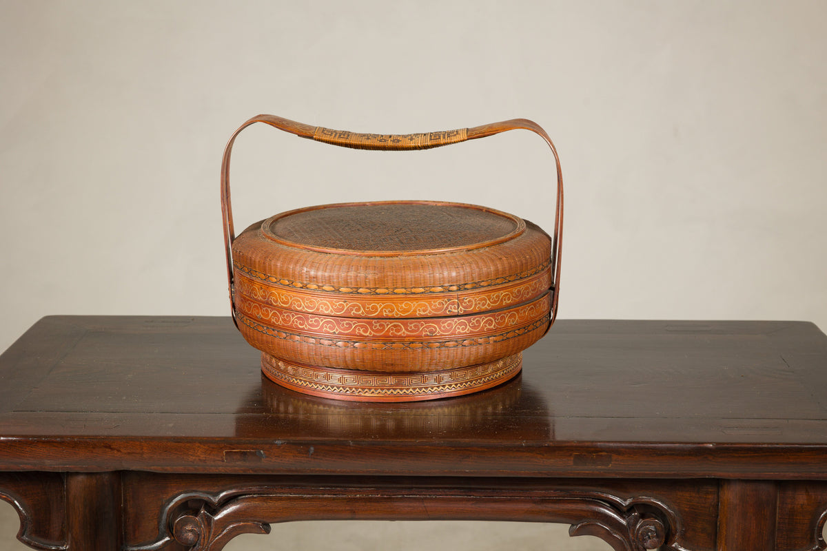 Brown woven rattan Chinese basket with painted cover and arched handle on a wooden table.