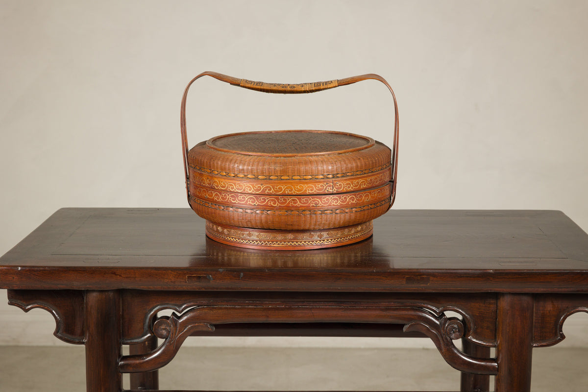 Brown woven rattan Chinese basket with painted cover and handle on a wood table.