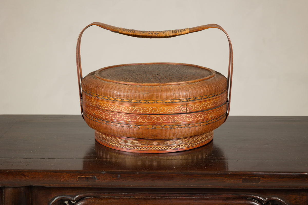 Gold and tan woven rattan Chinese basket with painted cover and arched handle on a table.