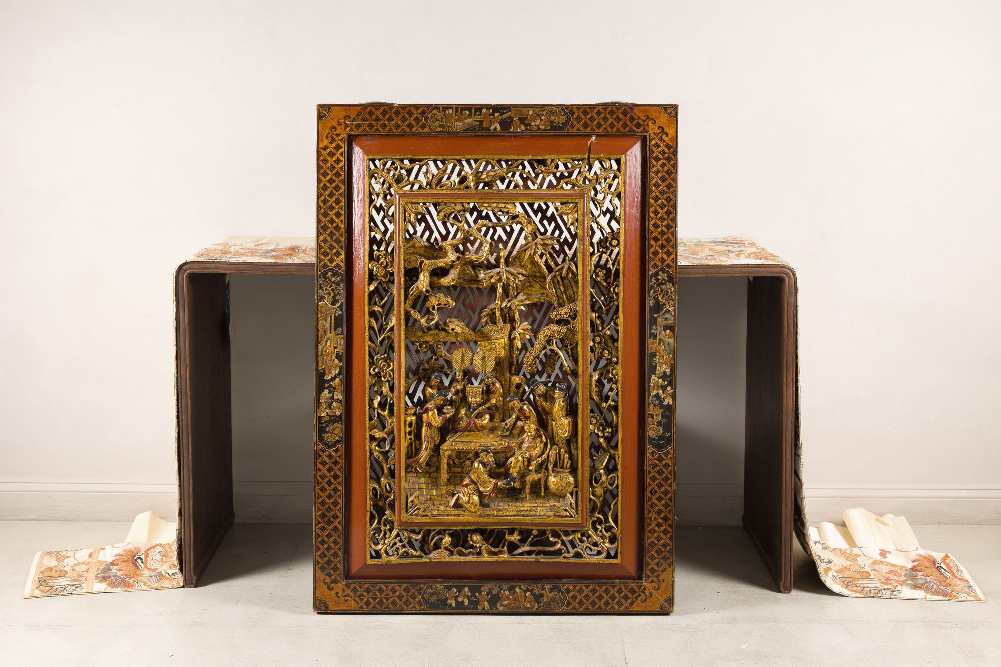 Gold giltwood Qing wall panel with figural court scene, displayed against patterned table and white wall.
