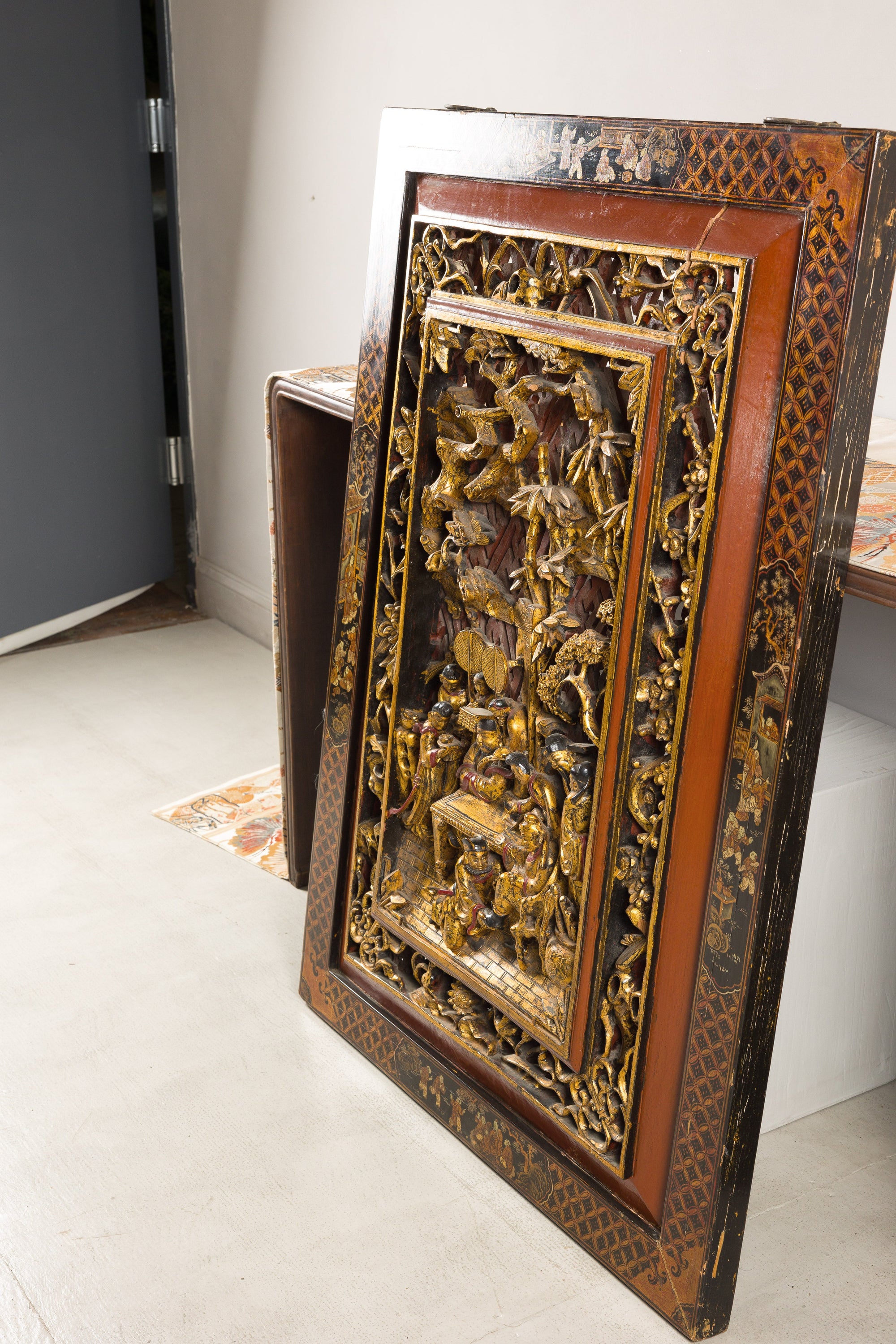 Gold giltwood Qing wall panel with carved figural court scene, leaning against a wall in a bright room.