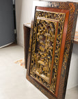 Gold giltwood Qing wall panel with carved figural court scene, leaning against a wall in a bright room.