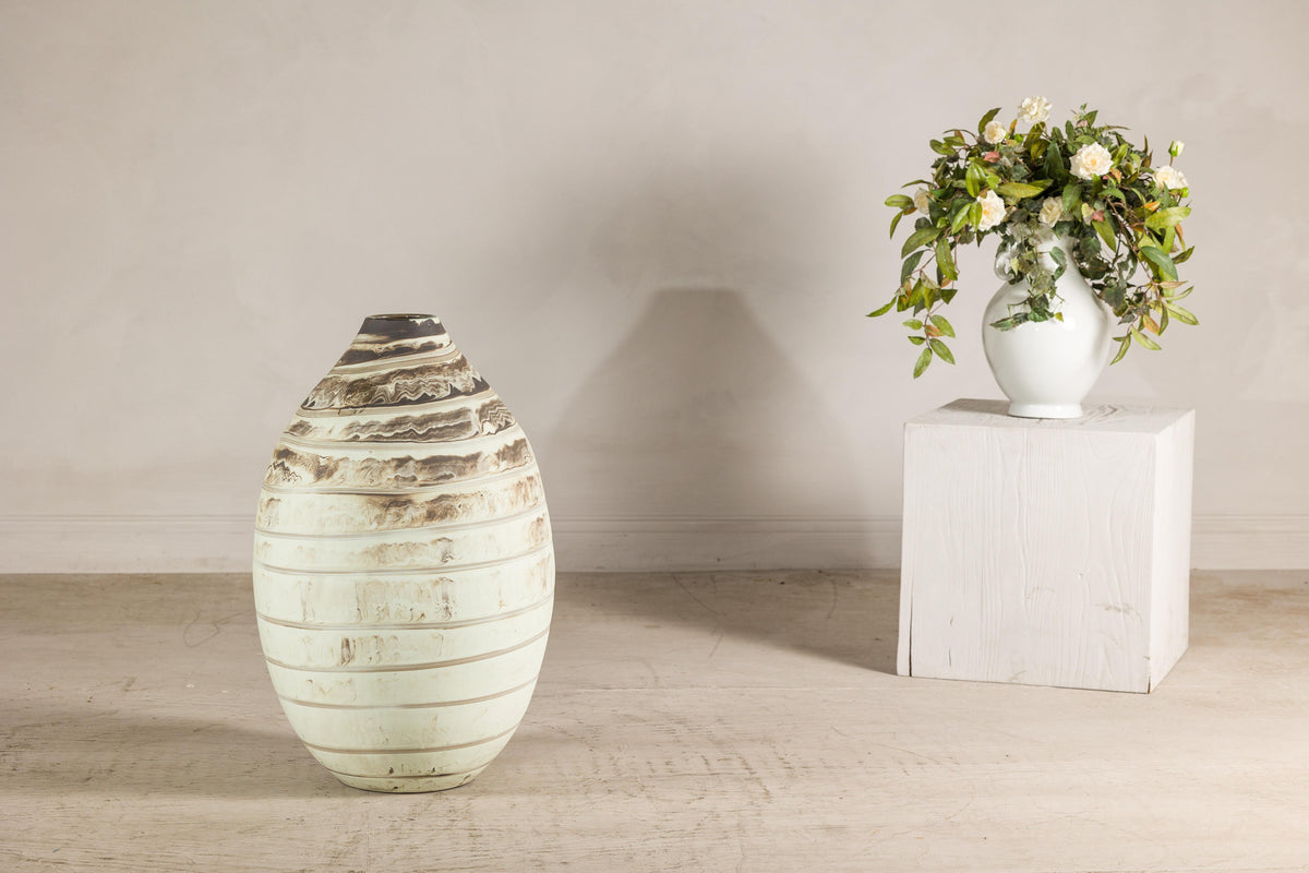 Cream-glazed artisan ceramic vase with brown bands on a floor next to a white pedestal with floral arrangement.