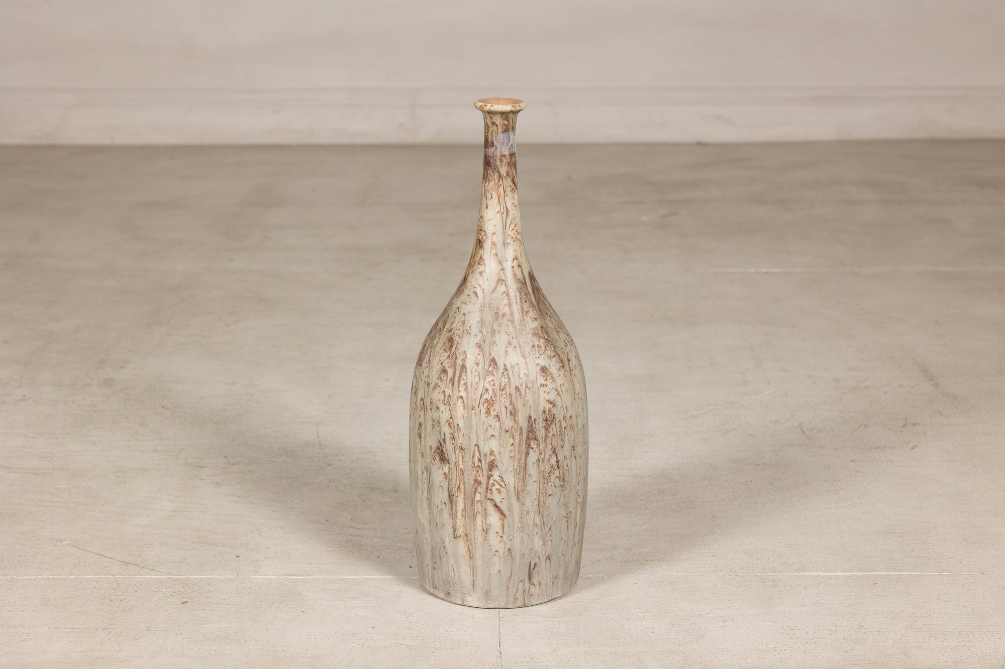 Cream and brown drip-glazed ceramic bottle vase on a plain light-colored floor, vertical view.