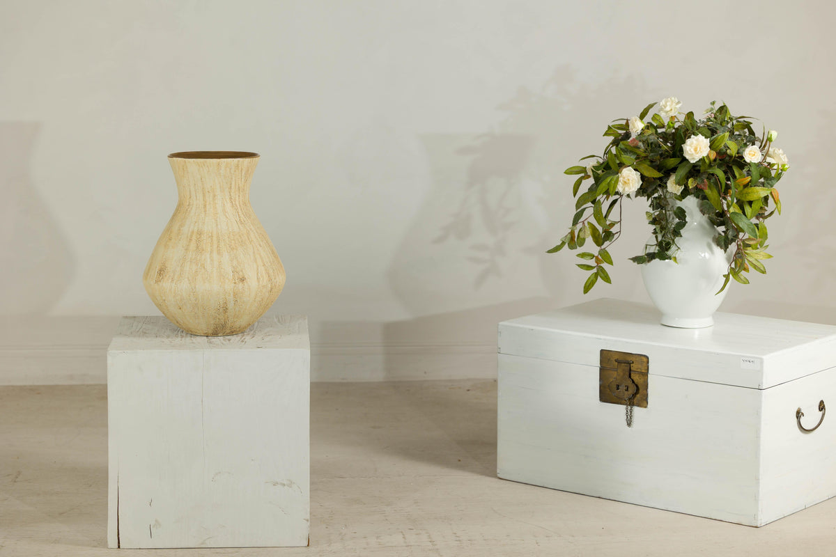 Drip glaze ceramic vase with flared neck on white block beside a flower-topped chest.