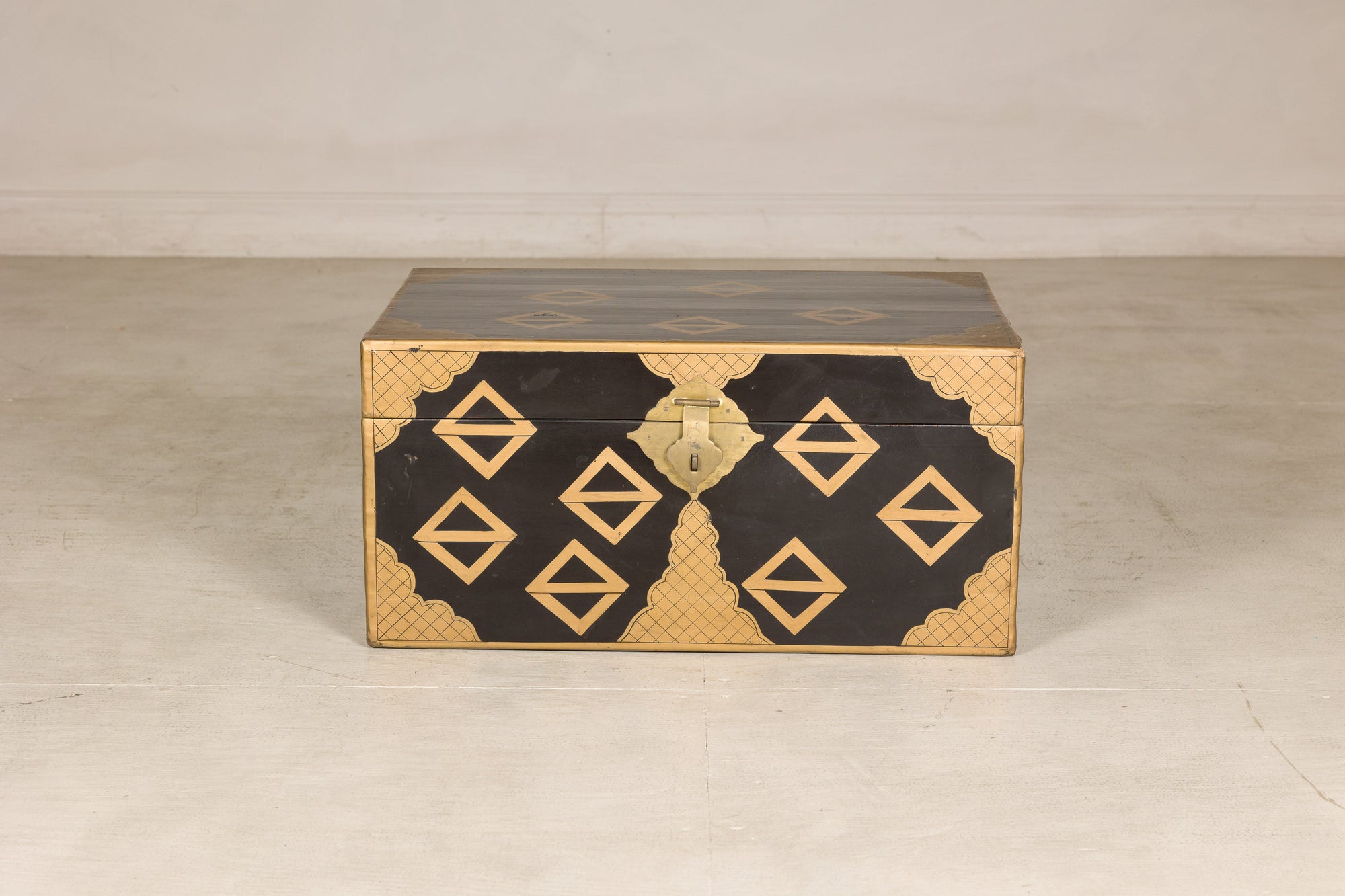 Black lacquered vintage Chinese chest with gold geometric decoration and central latch on a plain floor.
