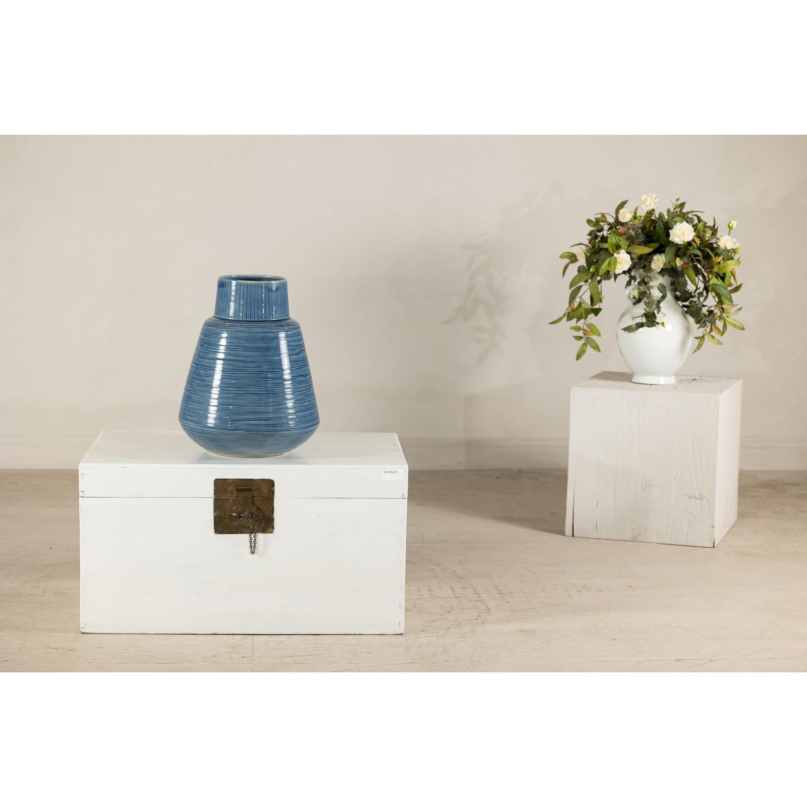Blue-glazed ceramic vase with tapering form on chest, white vase with flowers visible behind it.