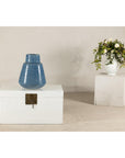 Blue-glazed ceramic vase with tapering form on chest, white vase with flowers visible behind it.