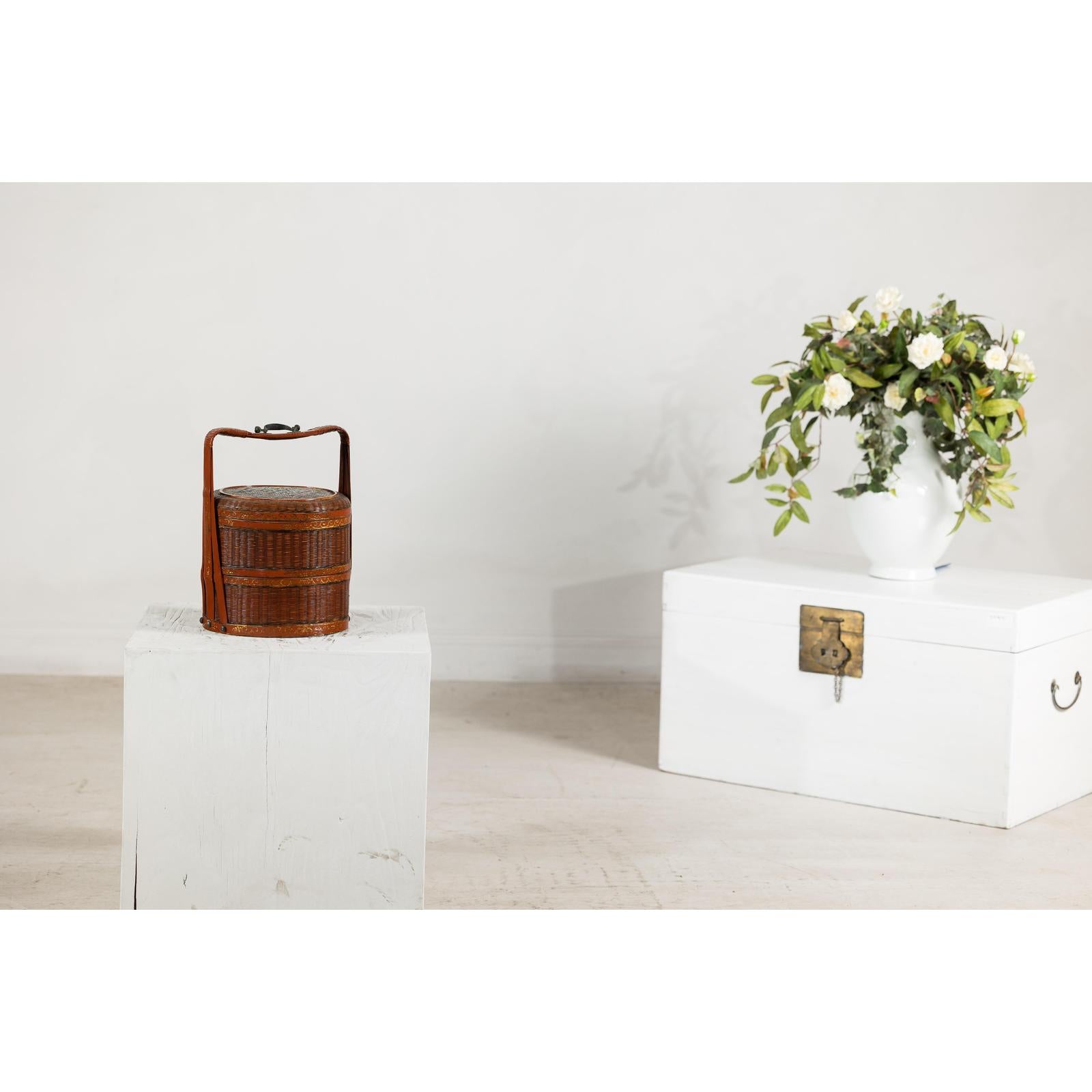 Brown vintage woven carrying box with double handles on white pedestal, next to a chest and vase of flowers.