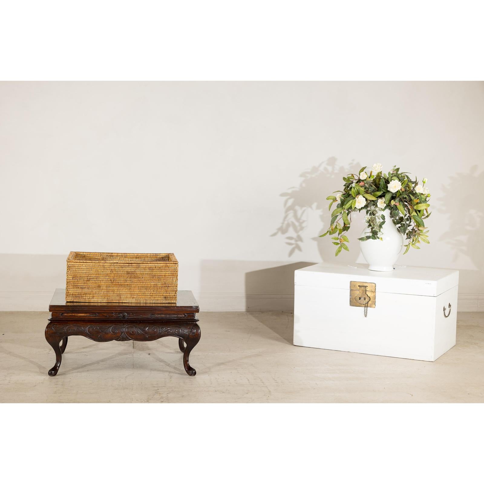 Brown rattan rectangular storage box with lid on wooden table beside a white trunk with flowers, country style setting.