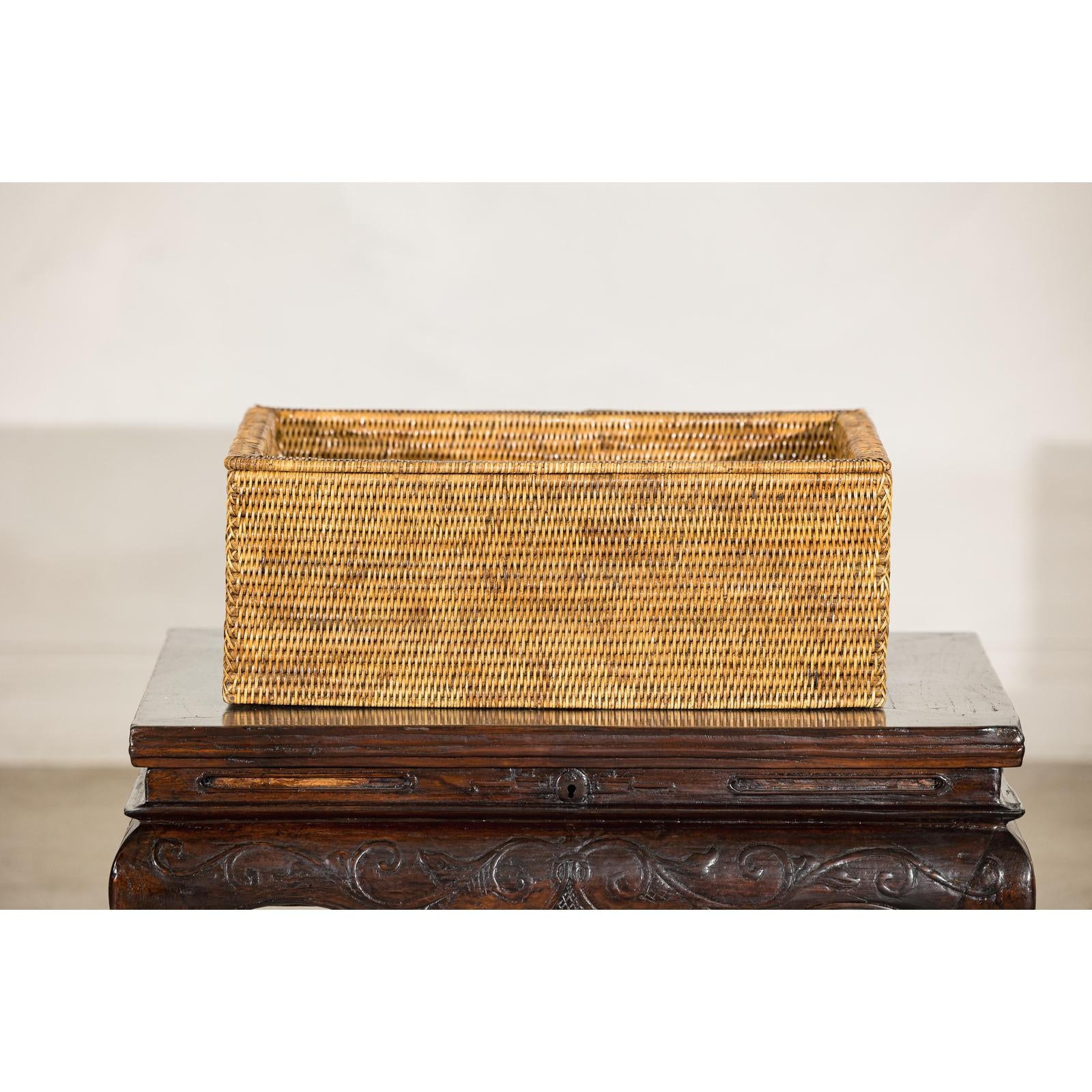 Brown rattan long rectangular storage box with lid on carved wooden table, plain background.