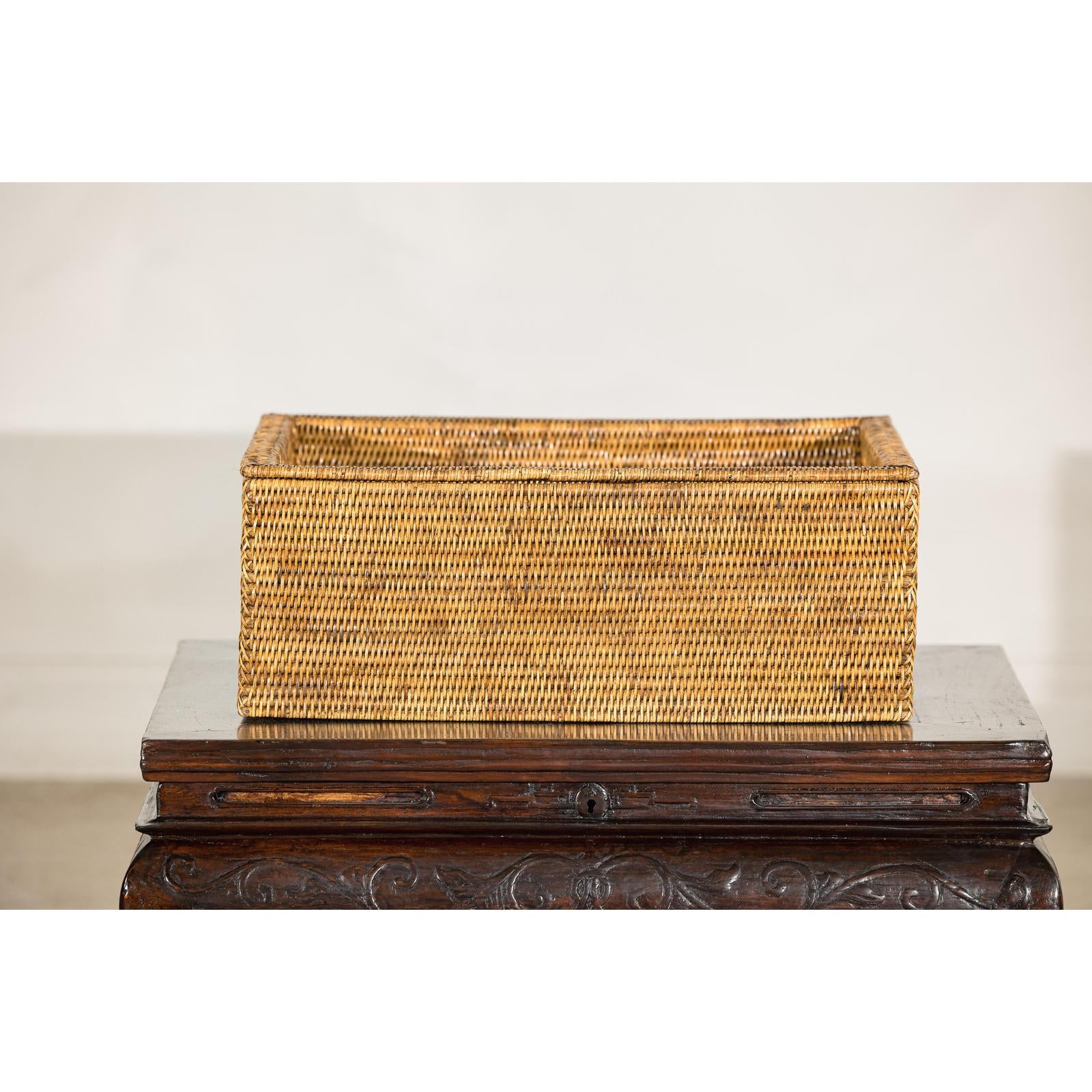 Brown rattan long rectangular storage box with lid on a dark carved wooden table against a plain background.