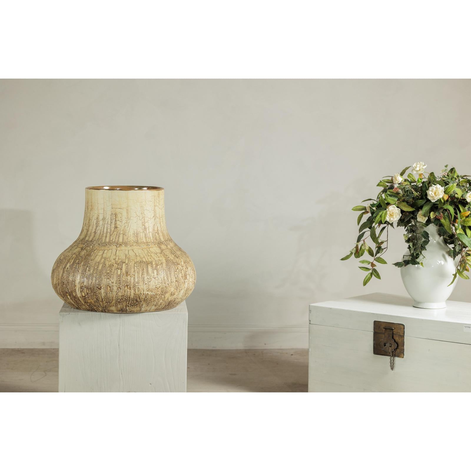 Cream and earth-toned textured ceramic vase on pedestal next to white chest with a flower vase in background.