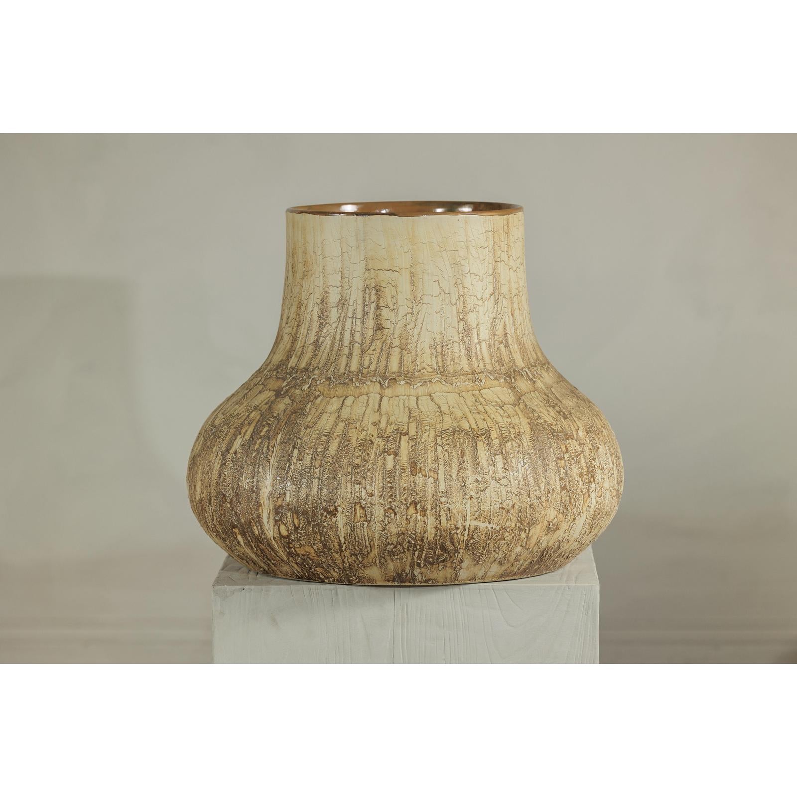 Textured cream and earth toned ceramic vase on white pedestal against plain background.