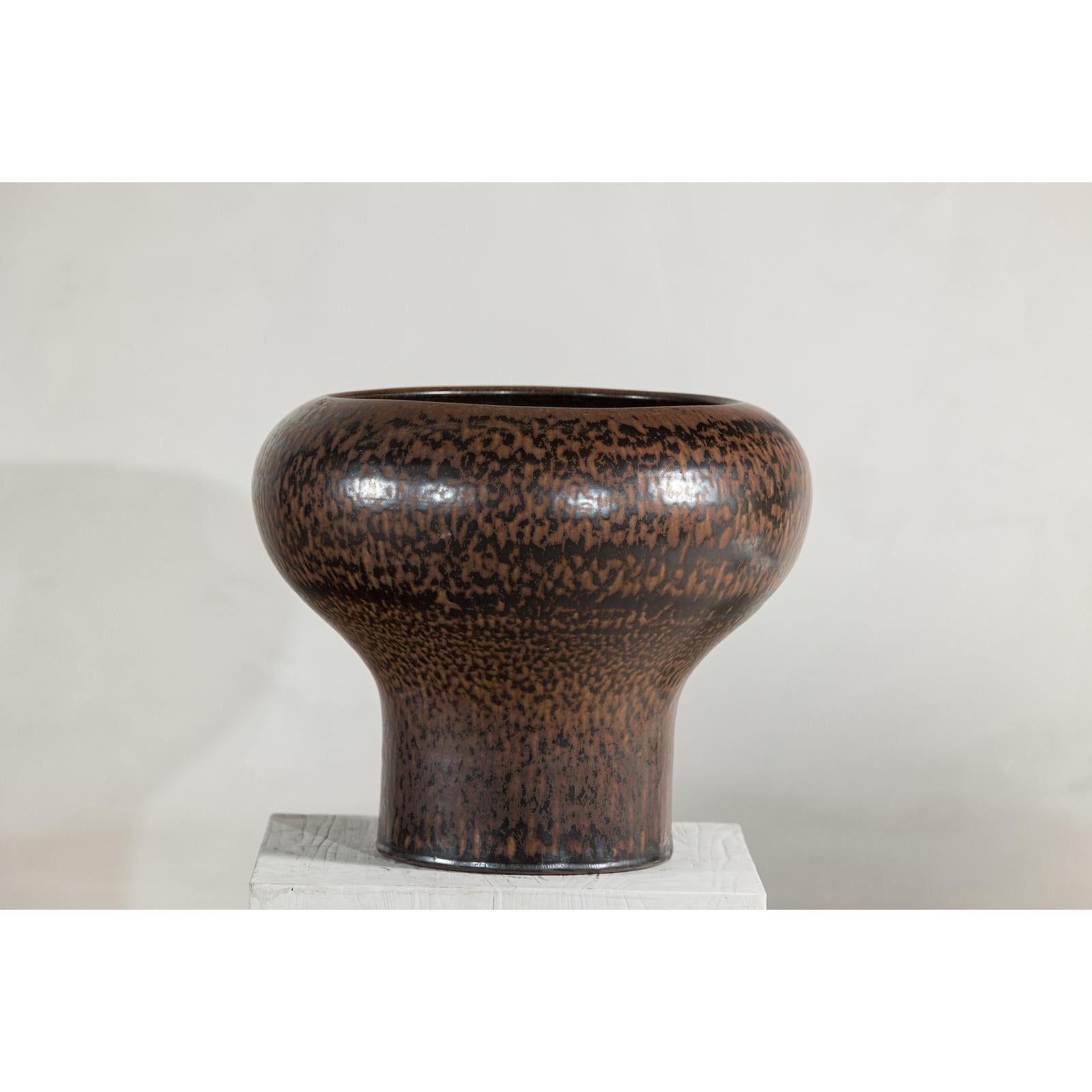 Speckled brown ceramic vase with wide mouth, rounded top, and narrow base on a white stand, close-up.