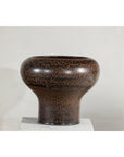 Speckled brown ceramic vase with wide mouth, rounded top, and narrow base on a white stand, close-up.
