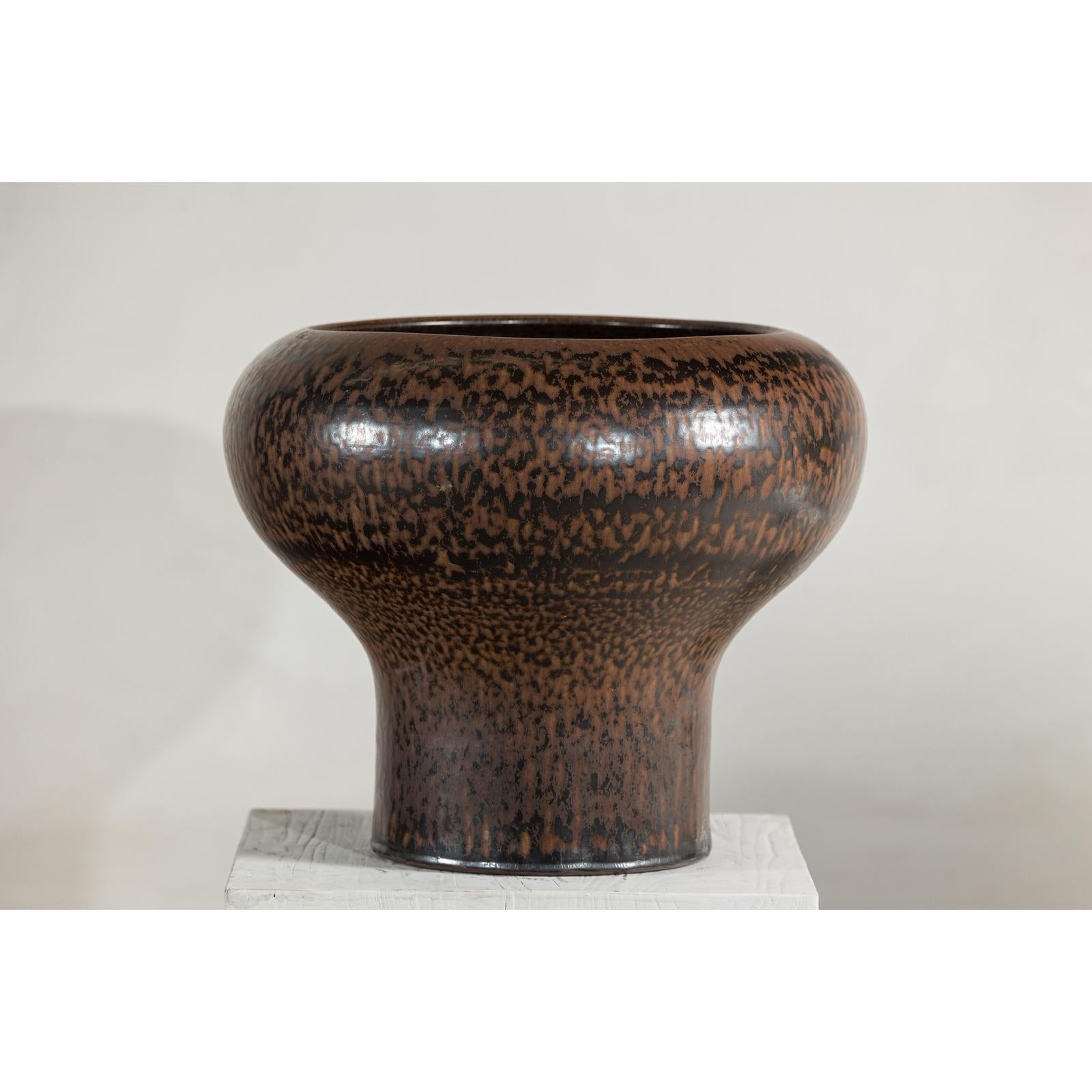 Speckled brown ceramic vase with wide mouth on white pedestal.