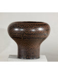 Speckled brown ceramic vase with wide mouth on white pedestal.