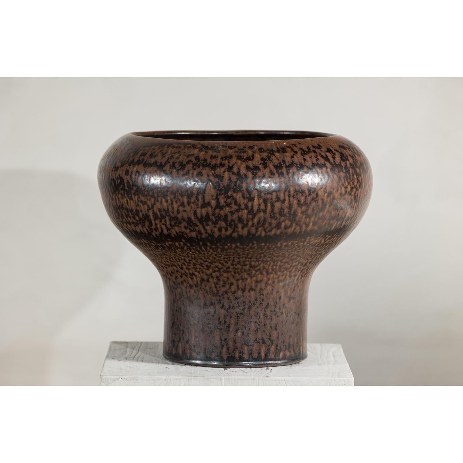 Brown speckled ceramic vase with wide mouth and narrow base on white pedestal, round shape, close-up view.