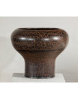 Brown speckled ceramic vase with wide mouth and narrow base on white pedestal, round shape, close-up view.