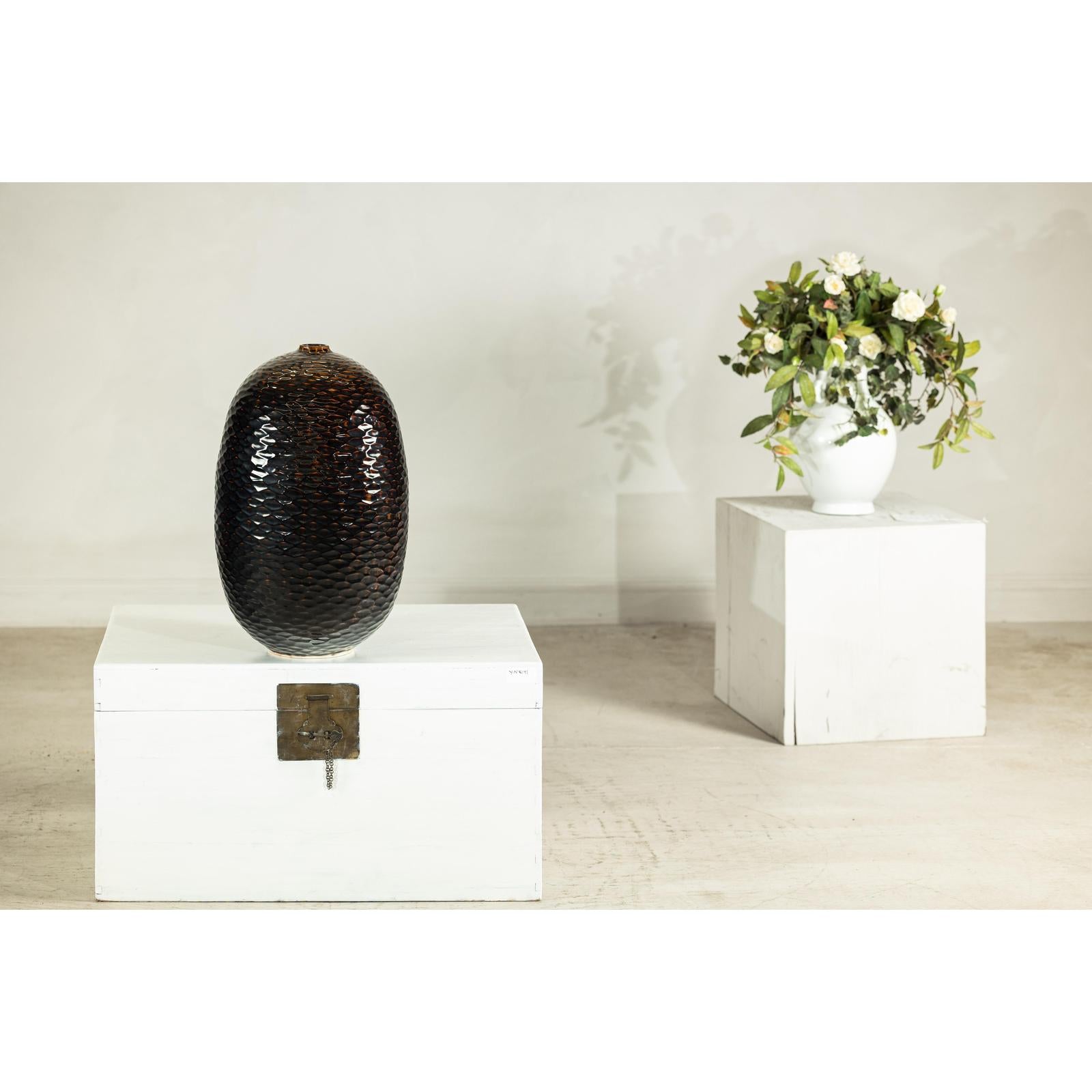 Deep brown and black glazed ceramic vase with honeycomb texture on white chest, white vase in background.