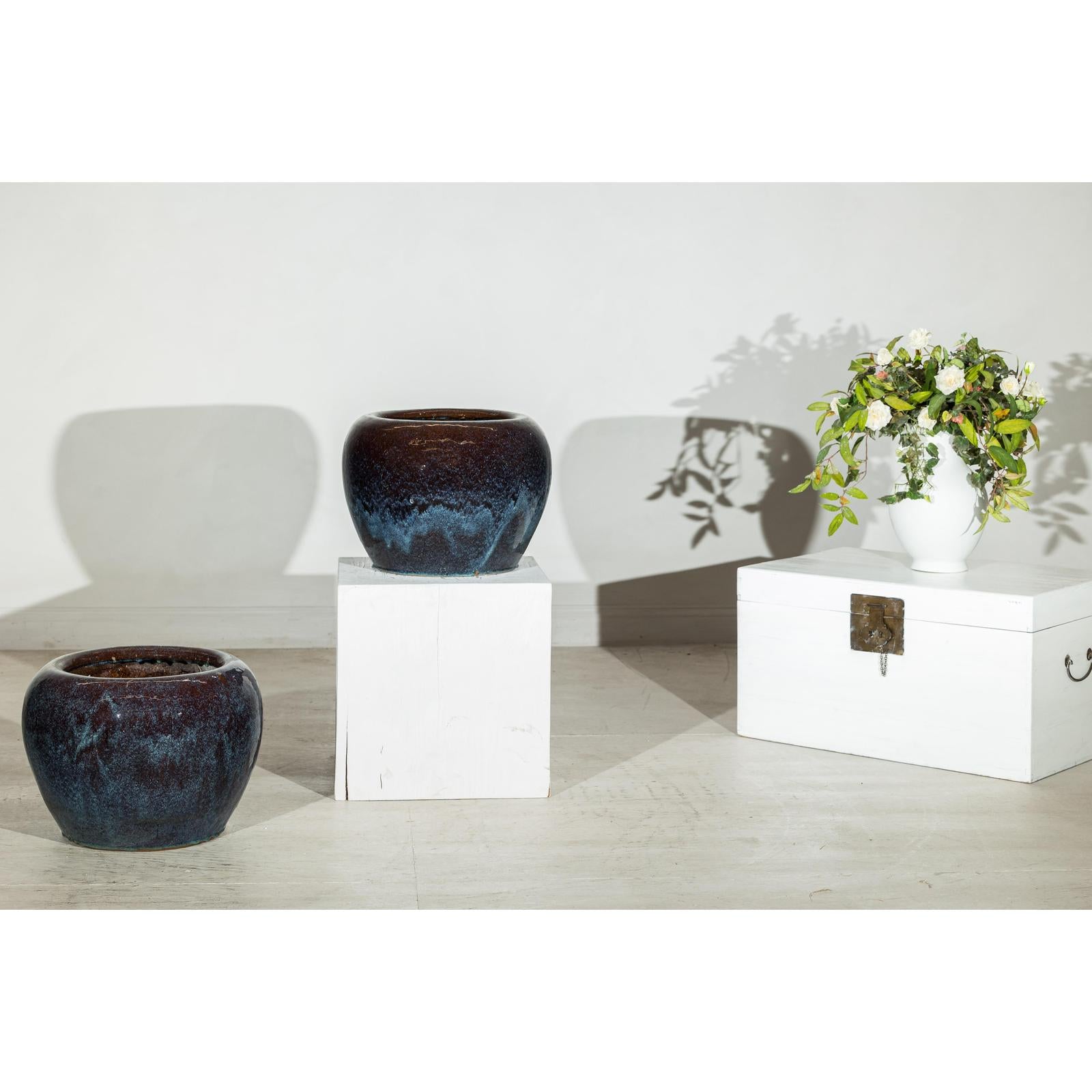 Close-up of two vintage blue and aqua glazed ceramic planters next to a white vase with flowers, shadows on wall.