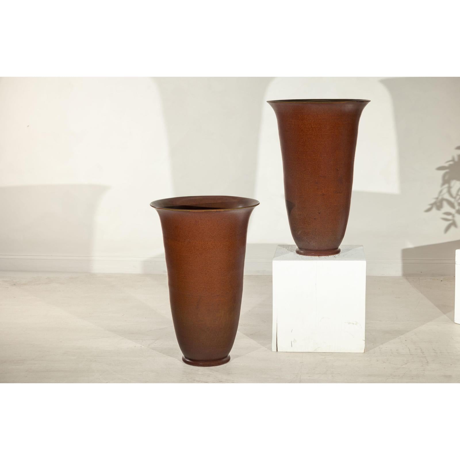 Brown ceramic vases with red undertones, one on the floor and one on a white pedestal against a light wall.