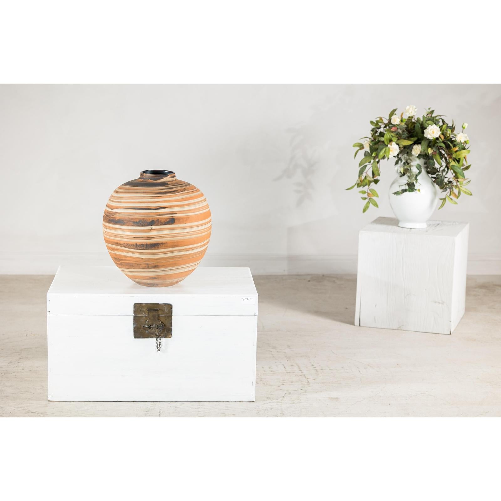 Round earth-tone ceramic vase with swirled design on white chest; white flowers on pedestal in background.