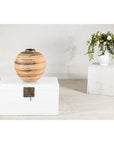 Round earth-tone ceramic vase with swirled design on white chest; white flowers on pedestal in background.