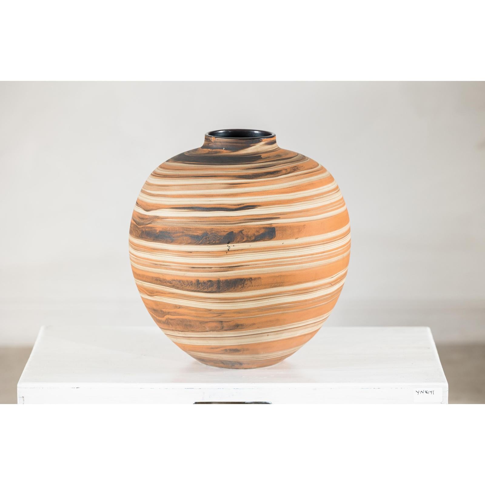 Close-up of a round ceramic artisan vase with swirled earth-tone design on a white surface against a plain background.