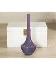 Purple ceramic vase with slender neck, artisan style, in front of a white wooden chest with brass latch.