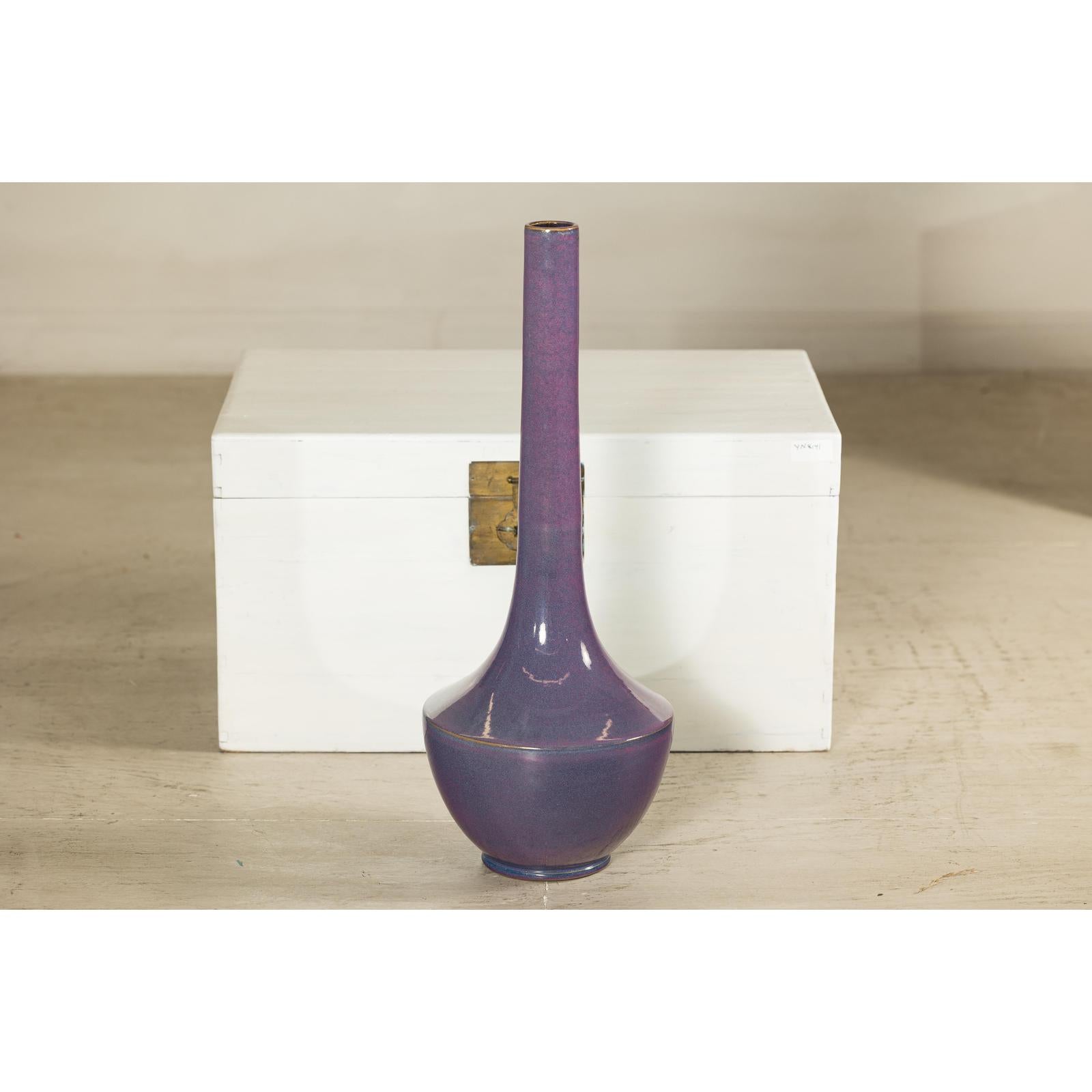 Purple ceramic vase with slender neck, artisan style, in front of a white chest on a beige floor.
