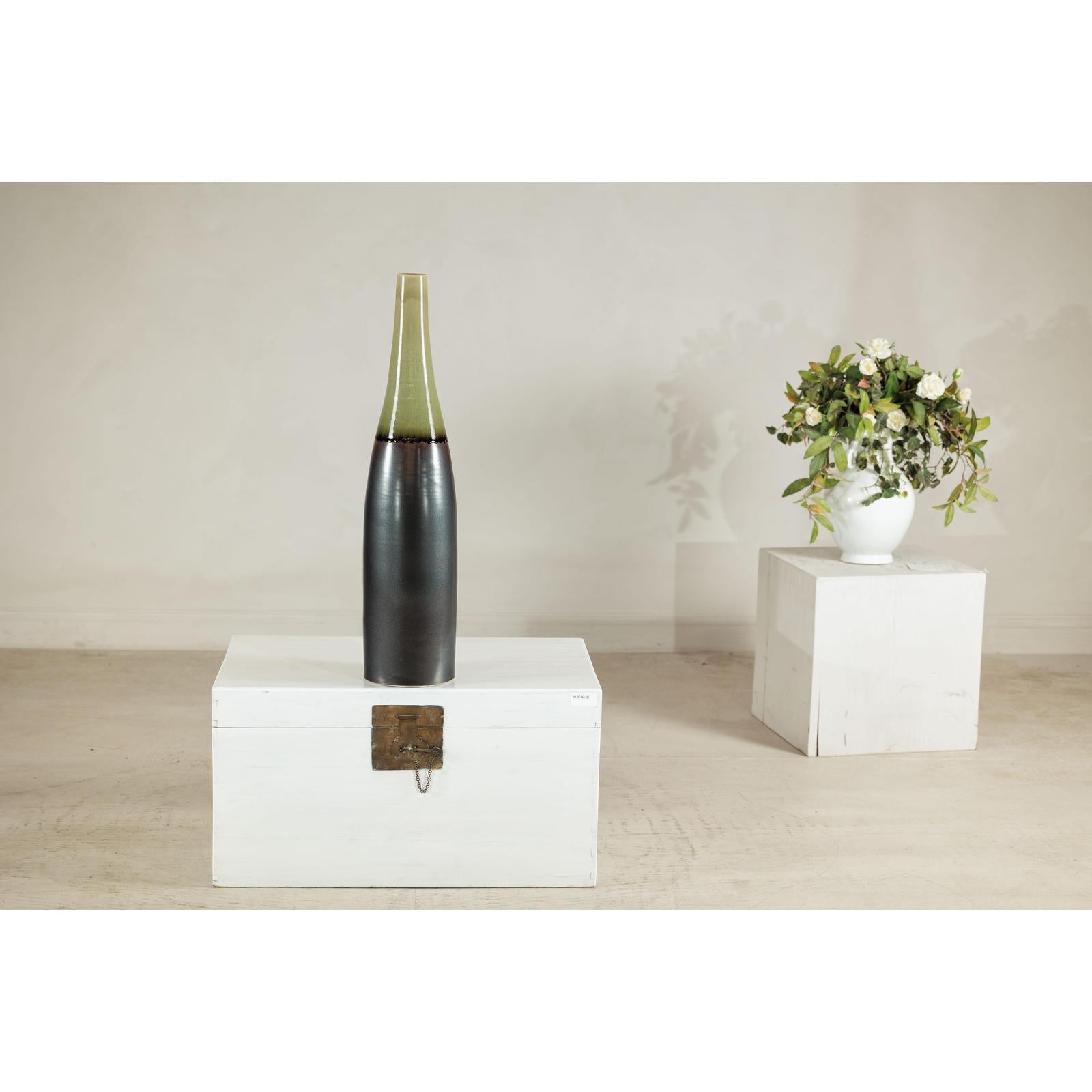 Charcoal ceramic vase with moss green glazed neck on white chest; white vase with green flowers in background.