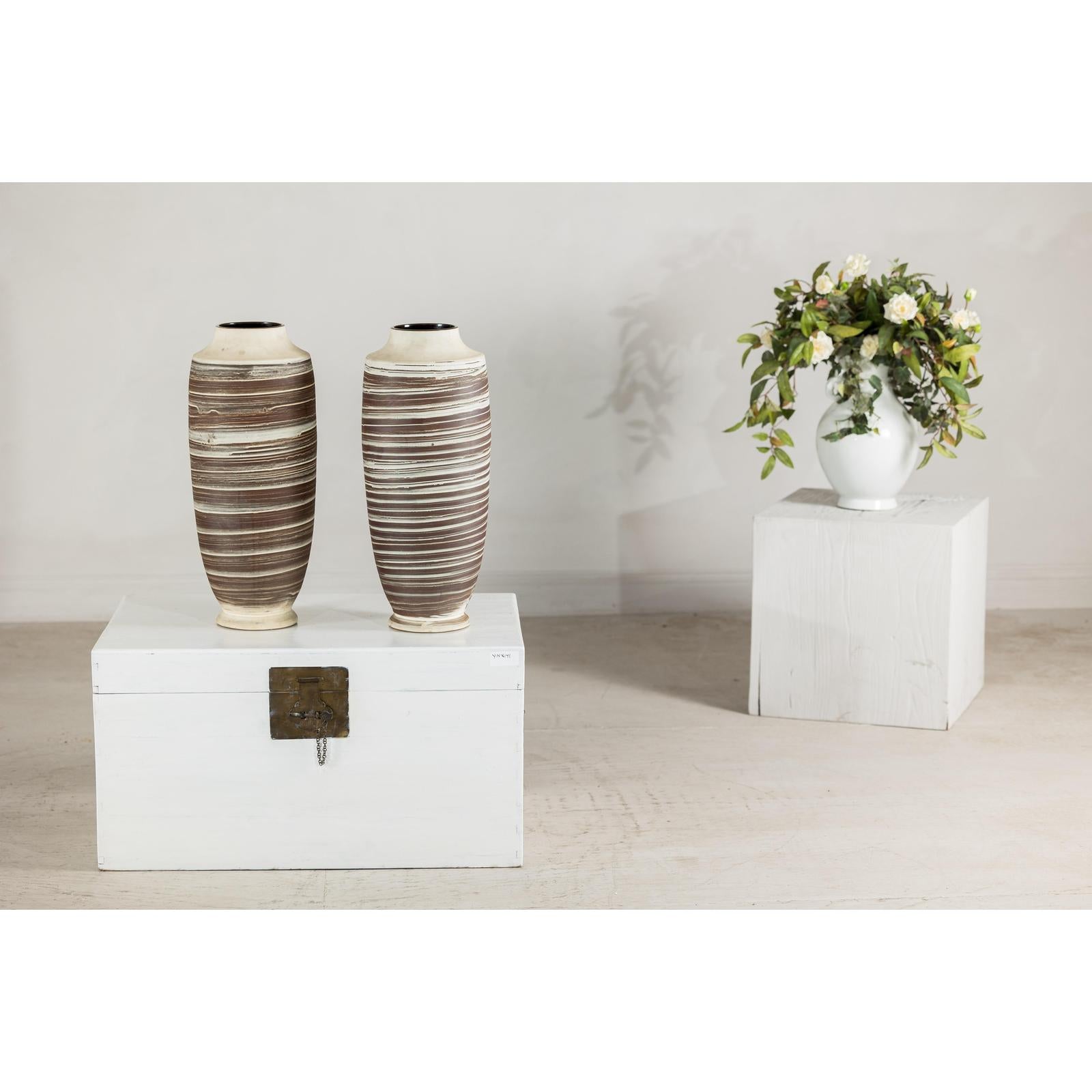 Close-up of two tall ceramic vases with horizontal swirl design on a white chest, flowers in white vase in background.