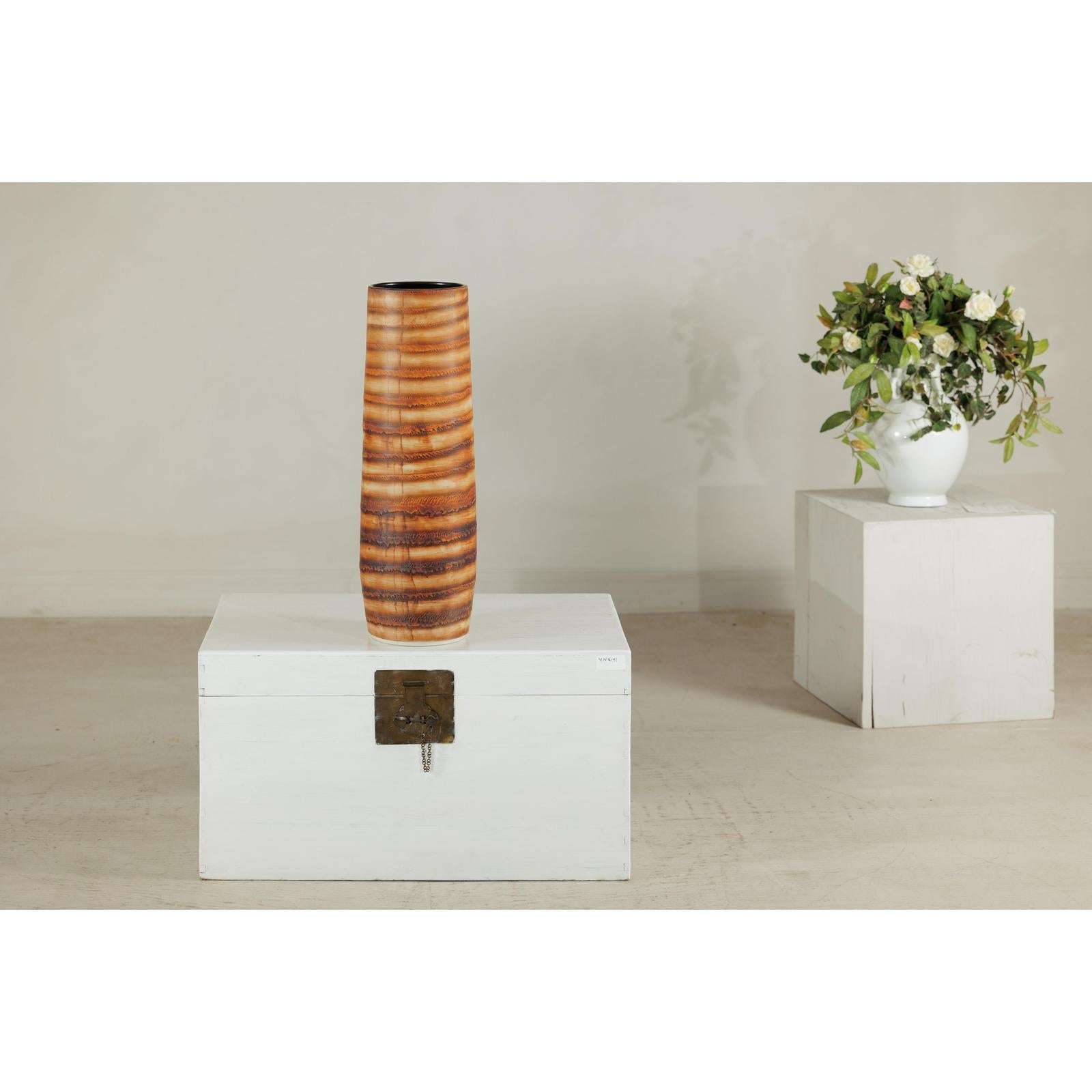 Orange-brown ceramic artisan vase with swirl design on white chest, white flower vase on pedestal in background.