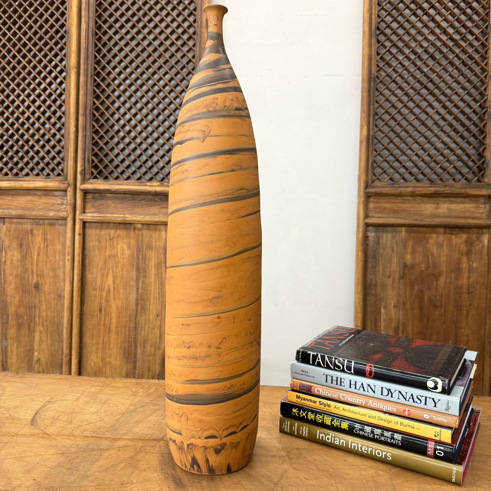 Orange vase with black swirl, ceramic, tall and skinny, next to books on a wooden table with a lattice cabinet behind.