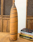 Orange vase with black swirl, ceramic, tall and skinny, next to books on a wooden table with a lattice cabinet behind.