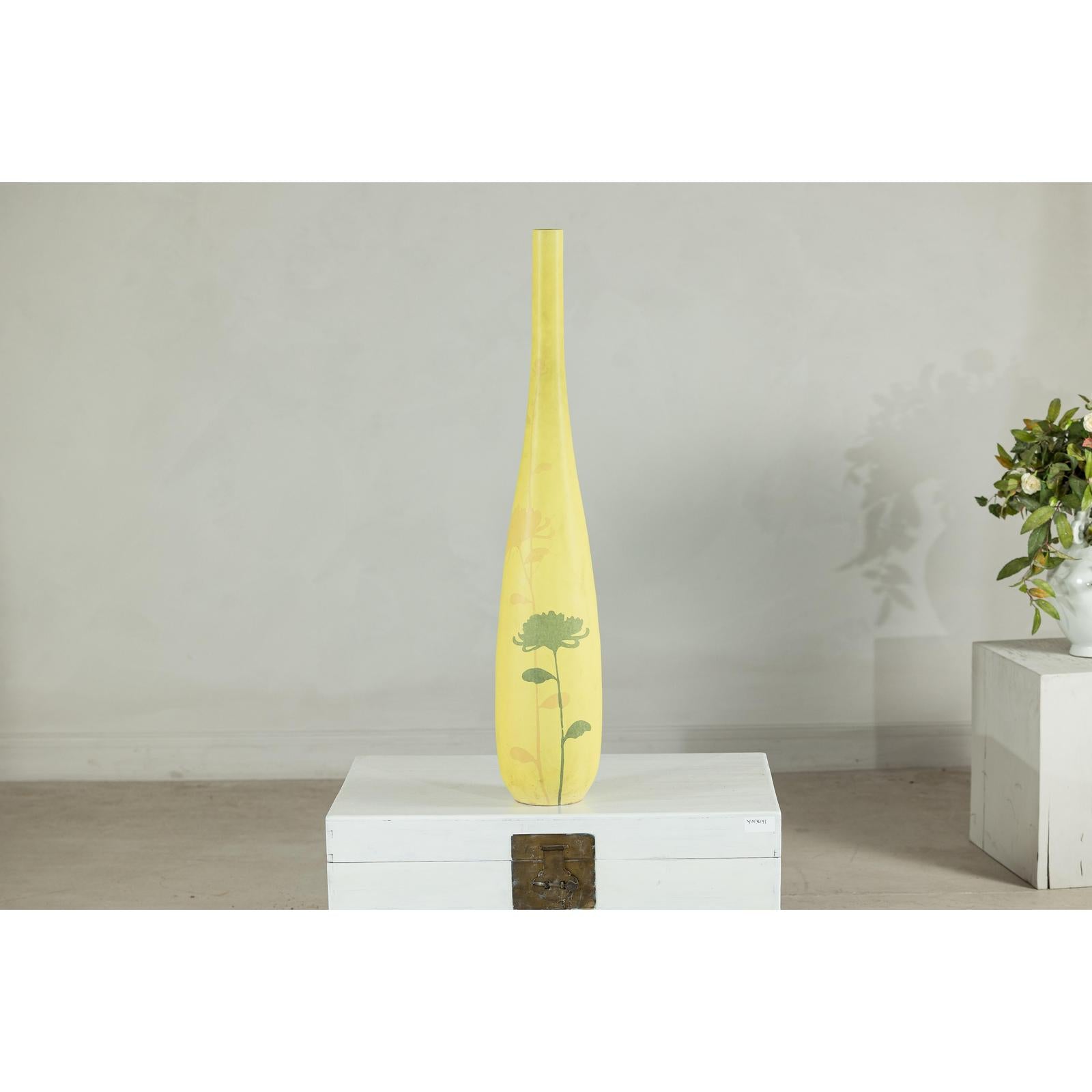 Yellow ceramic vase with hand-painted flowers, tall and skinny, on white pedestal against a plain wall.
