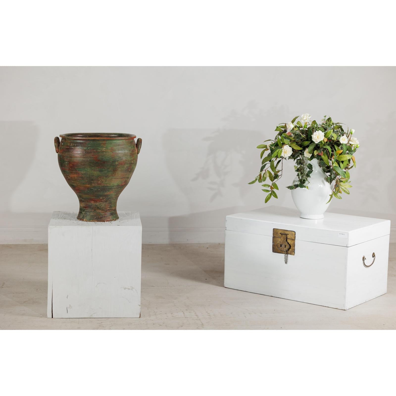 Green, brown, and red vintage ceramic planter on a white pedestal next to a chest with flowers on top.