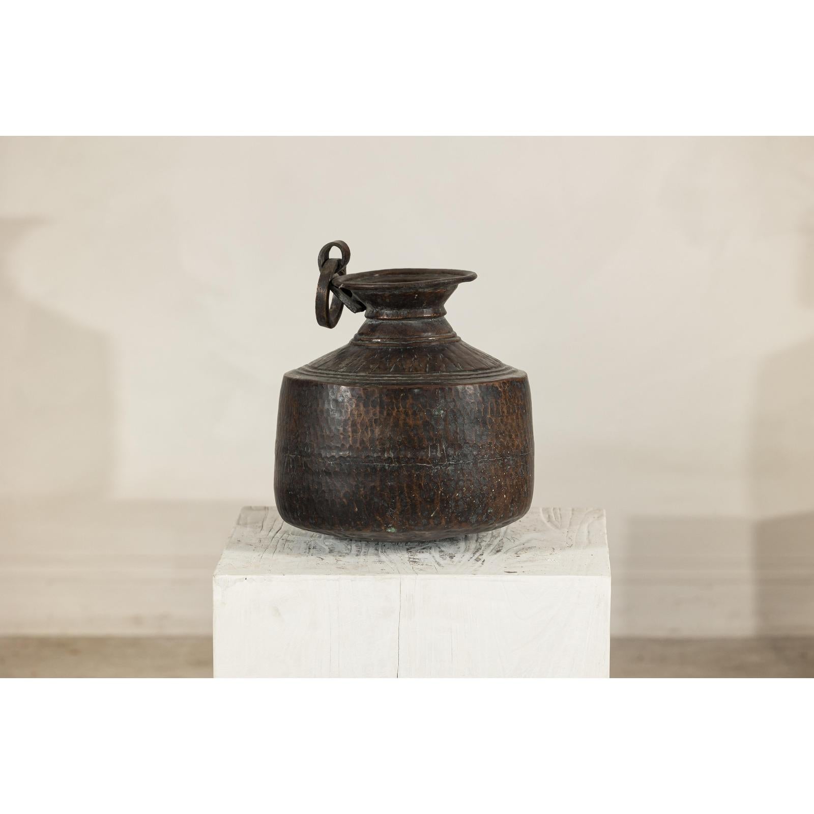 Close-up of a vintage hand-hammered metal jug with ring handle on a white pedestal against a plain background.
