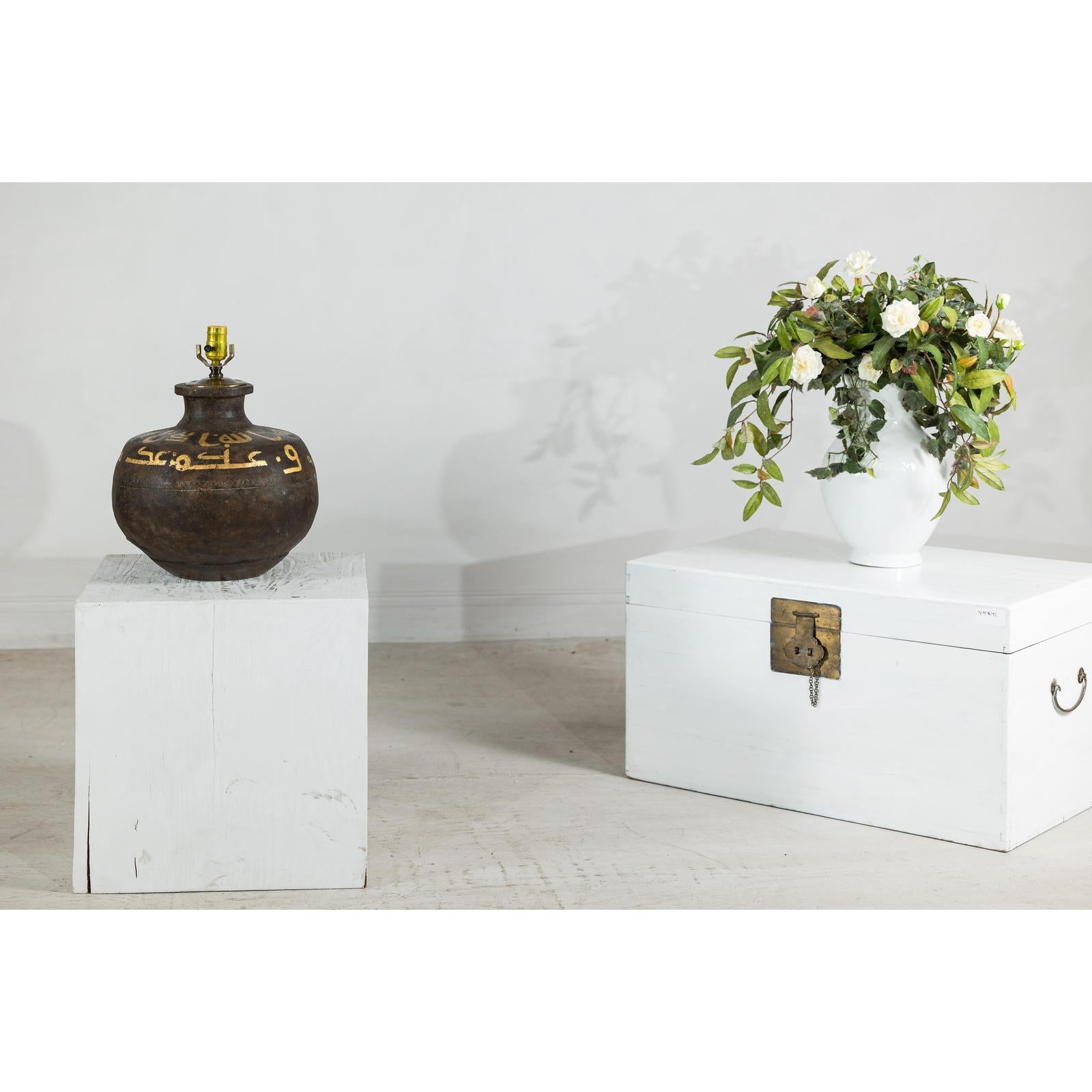 Close-up of a vintage metal jug lamp with painted script next to a white vase with flowers on white pedestals by a chest.