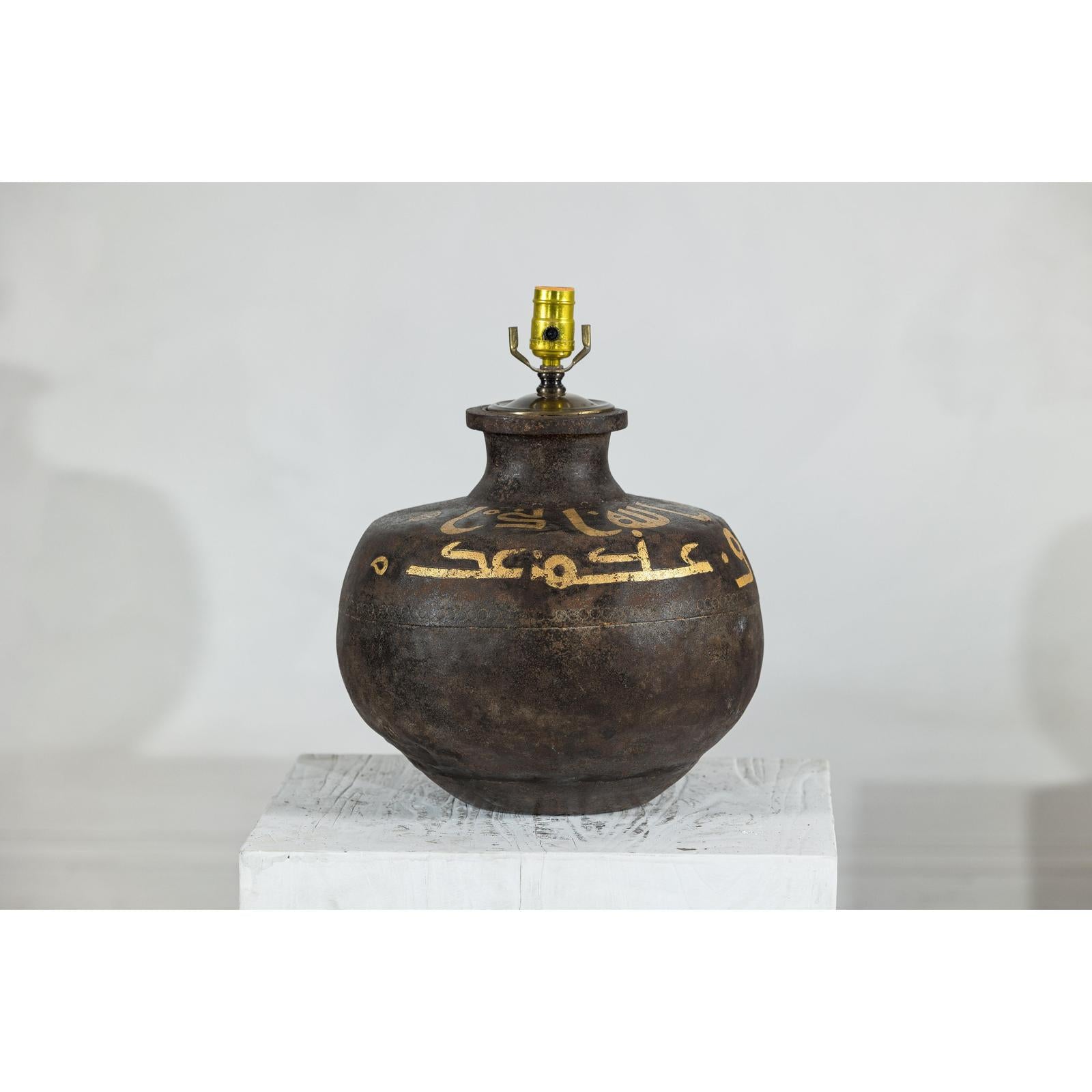 Close-up of a vintage metal jug lamp with painted script on a white pedestal.